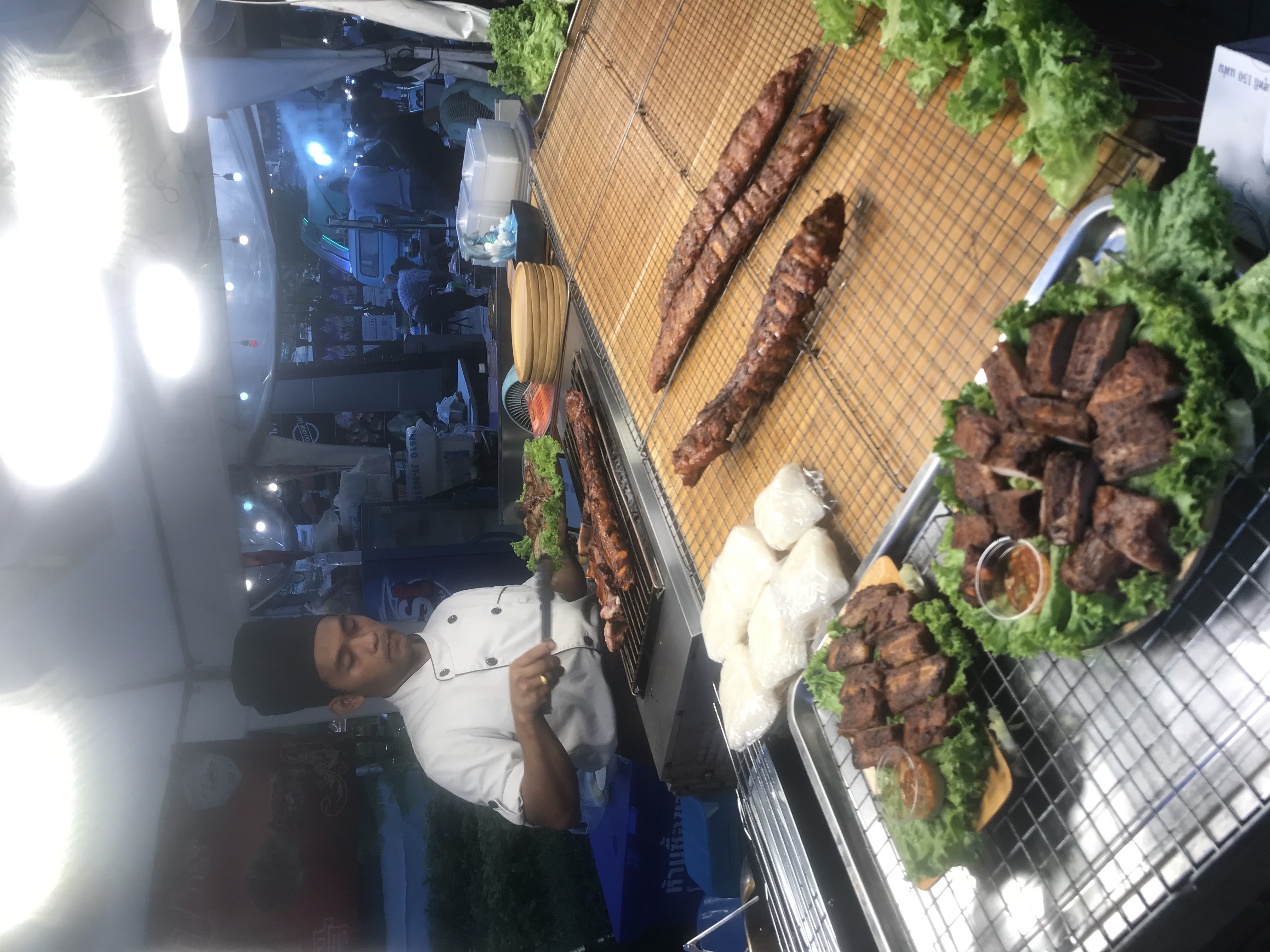 Bangkok night market — chef working a live BBQ grill, long skewers of grilled meat and platters of ribs on lettuce